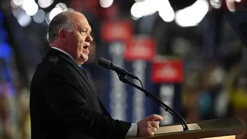 Getty Images Tom Homan speaks into a microphone in front of a lectern. He wears a black suit jacket and white shirt.