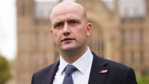 pa Stephen Flynn, a bald man wearing a dark blue suit and tie with a white shirt, stands in front of the Houses of Parliament 