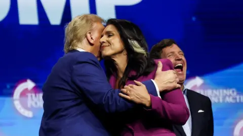 Reuters Trump and Gabbard embrace on stage in Duluth, Georgia, on campaign trail in October, with Tucker Carlson in background laughing