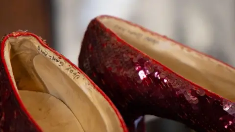 Heritage Auctions A close-up of the ruby red sequin shoes which feature dark red sequins sewn in diagonal strips across the side of the shoes, with a light coloured insole. The name 'Judy Garland' is written in capitals in black pen on the inside seam of each shoe