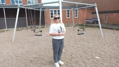 A woman with blonde hair and sunglasses stands in front of a bare playground made of sand and run-down swings, in front of an old school building.