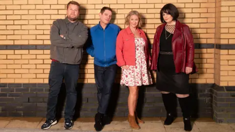 BBC/Fulwell73 & Tidy Productions/Tom Jackson Four members of the cast of Gavin and Stacey reunite for an official cast photo: Smithy, Gavin, Stacey and Nessa. They are stood against a brick wall. Smithy (played by James Corden) is wearing a grey jacket and blue jeans and has his arms crossed, Gavin (played by Matthew Horne) is next to him, wearing a blue jacket with his hands in the pockets and blue jeans. Next to him is Stacey (played by Joanna Page), she is wearing brown cowboy boots, a white floral dress and a pink jacket, and she is stood next to Nessa (played by Ruth Jones), who is wearing a burgundy leather jacket and a black dress with long black boots. 