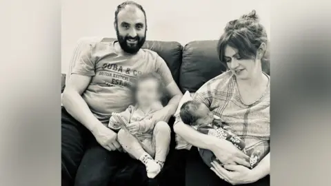 Family handout A man and a woman sitting on a sofa with two children. The man has dark hair with a receding hairline and a dark beard. He is smiling at the camera and has his arms around a toddler who has their face blurred. The woman is looking down at a sleeping baby in her arms.