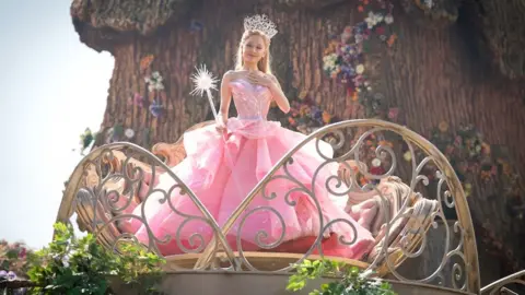 Universal Ariana Grande wears a pink gown complete with a crystal embellished crown and wand.