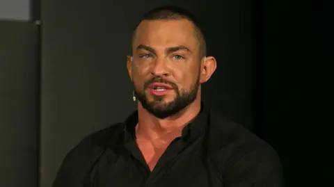 PA Robin Windsor wearing a collared black shirt. He has an earpiece and a beard. He is looking directly at the camera.