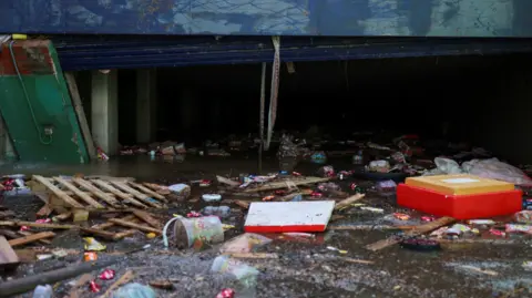 Reuters Brown water can be seen flooding the entrance of a car park, with debris including wooden pallets and plastic packaging floating in it.