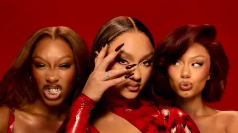 Tom Emmerson Promotional photo for the girl group Flo, showing all three members against a crimson red background. Singer Reneé Downer bares her teeth, bandmate Stella Quaresma poses with her hand partially obscuring her face, and Jorja Douglas pouts for the camera.