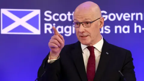 Getty Images John Swinney gives a speech in Edinburgh. He is wearing a black suit jacket, a maroon tie and a white shirt.