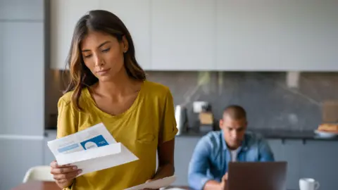 Getty Images Woman looking at a bill in the kitchen of a home with a man sitting in front of a laptop in the background.