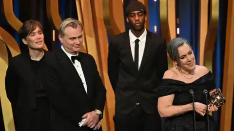 Getty Images Cillian Murphy, Christopher Nolan, and Emma Thomas accept the best film award for Oppenheimer on stage during the Bafta Film Awards 2024 at the Royal Festival Hall on February 18 in London, while a YouTube prankster stands in the background