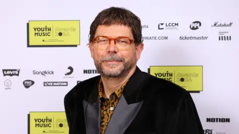 Youth Music Matt Griffiths at a Youth Music event. He wears a black velvet jacket over a leopard-print shirt, paired with round orange and yellow ombre glasses. 