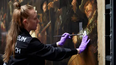 Courtesy of the Rijksmuseum A woman with long blonde hair in a ponytail is wearing black protective clothing and purple latex gloves as she carefully removes the varnish from a section of The Night Watch