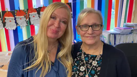 BBC A woman with blond hair and a blue top stood beside an older lady with grey hair and black framed glasses in a dark top with flowers on. Both are stood in front of a multi coloured striped wall