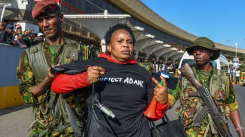 Getty Images At the airport, two armed soldiers in military uniforms escort a female protester holding a vuvuzela and dressed in a black T-shirt that says 