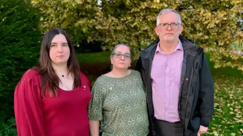 Steve Hubbard/BBC Declan's sister Kaitlyn, mother Sam and father Graeme, standing together in front of trees shedding their leaves. Kaitlyn is in her 30s and is wearing a red jumper, while Declan's mother is wearing a patterned green top and has blue rimmed glasses. His father has grey hair, glasses and a light beard and is wearing a black rain jacket, a purple shirt and grey suit trousers. They are looking at the camera seriously.