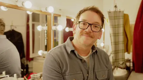 Steven Meo in his dressing room at the Apollo Theatre in London wearing glasses and a grey shirt, smiles at the camera