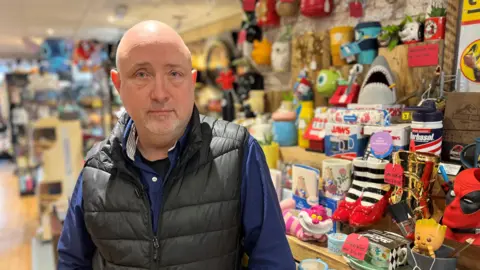 BBC Martin Gaunt, aged 62, wearing a blue shirt and black padded gillet, with colourful stock on shelves in the background.