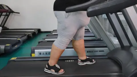 Getty Images An overweight woman wearing grey shorts and a black t-shirt walks on a treadmill in a gym.