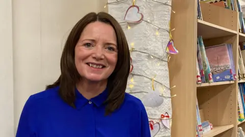 A woman with dark hair and a blue top in a school library with books on the wall and some Christmas lights running along the wall