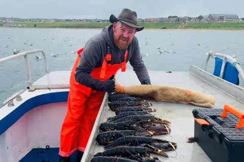 Richard Donaldson Man on boat wearing green cowboy-style hat, grey top and orange overalls, with several dark lobsters with their claws tied together.