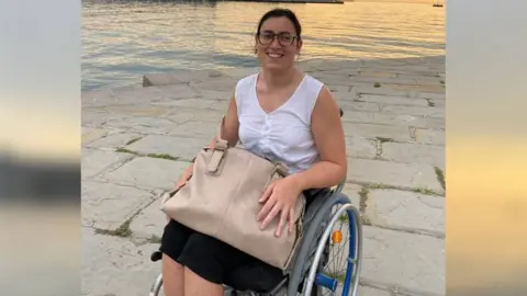 Sofia Brizio A woman in her twenties sits in a wheelchair with a large bag on her lap next to water, she smiles and looks towards the camera