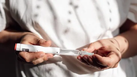 Getty A woman wearing a white t-shirt holds a white injectable medicine pen in front of her stomach