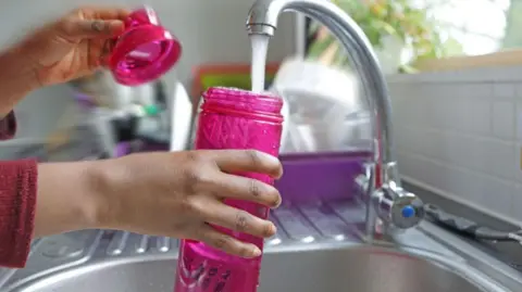 Getty Images A woman fills up a pink bottle with water from a kitchen tap in a bright kitchen.