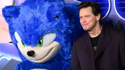 Getty Images A photo taken at a film premiere, sees Jim Carrey standing beside a person in a giant fluffy blue Sonic the Hedgehog costume.