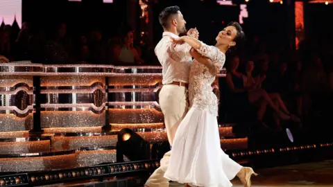 PA Media Both dressed in white and cream colours, Giovanni Pernice and Amanda Abbington dance on the Strictly stage. The judges' panel can be seen behind them as well as some audience members although the light is focussed on the couple. 