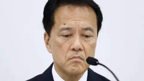 Getty Images Kentaro Okuda wearing a suit looks down with a microphone in front of him