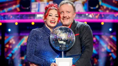 BBC Dianne Buswell and Chris McCausland standing together, smiling and holding the glitterball trophy after winning the Strictly Come Dancing final