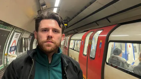 BBC Niall McNamee, standing on a London underground platform while a tube passes on the left-hand side. Niall has dark, short hair, and a beard. He is wearing a black jacket, with a green fleece underneath.