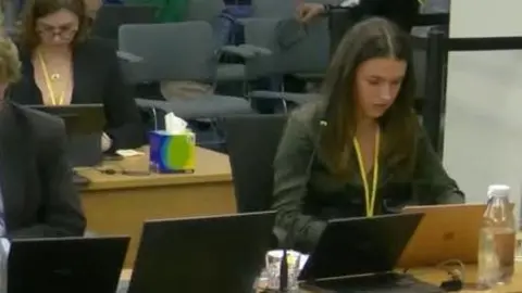 Post Office Inquiry Isabella Thomas sitting at the inquiry with a yellow lanyard round her neck looking at her laptop, with other laptops seen on the desk next to her