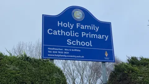 A blue school sign with the words Holy Family Catholic Primary School written in white lettering and green hedges behind it