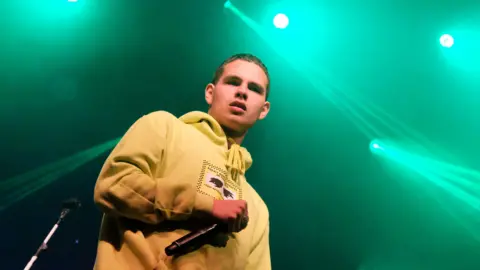 BBC Slowthai wearing a yellow hoodie and holding a black mic. He has short brown hair. There are green lights behind him.