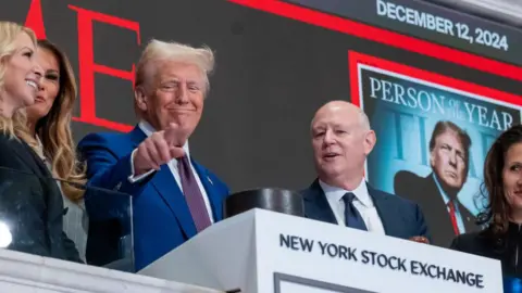 Getty Images President-elect Donald Trump is seen standing above the New York Stock Exchange (NYSE) on December 12, 2024 in New York City with his wife, Melania, partially visible and a few others around him. He is pointing and a screen showing the Time magazine cover with him as Person of the Year is behind him. 