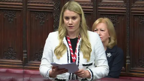 UK Parliament Baroness Owen speaking in the House of Lords chamber. She is holding sheets of paper and wearing a white jacket.