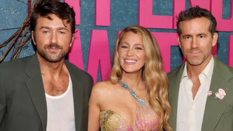 Getty Images Three people - Brandon Sklenar, Blake Lively and Ryan Reynolds - are dressed in formal attire and posing for a photo. They are stood in front of a large sign that reads 