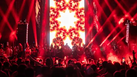 Siin experience Red, black and white lights glow behind a DJ and a crowd on and in front of a the stage at an electronic music concert in Syria 