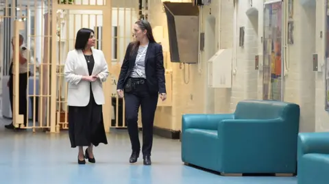 PA Media Shabana Mahmood walks with HMP Bedford Governor Sarah Bott, inside Bedford prison. Bars are behind them and they walk past a telephone and an armchair