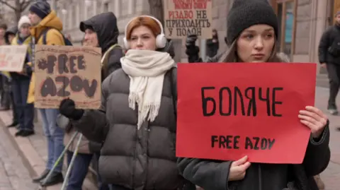 People are seen at a protest, including a young woman holding a red sign saying 