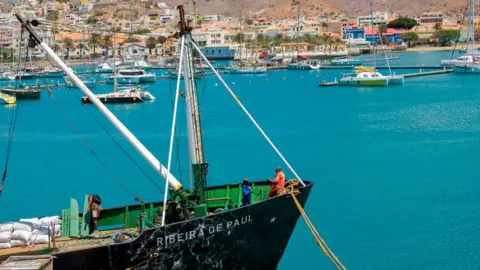 Getty Images Workers load goods on to a ship in Mindelo harbour in Cape Verde. Yachts can been seen in the background. The aquamarine sea is calm. 