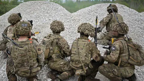 Getty Images Six soldiers wearing camouflage face away from the camera, kneeling in pile of grey stones while on exercise