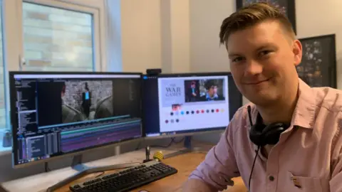 Rich Tipple Rich Tipple smiling to camera wearing a pink shirt with two large monitors behind him showing a scene from the colour version of The War Games