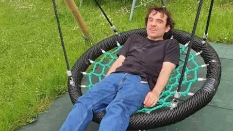 Family handout Declan Morrison sitting on a large netted, basket style swing. He is mid swing and is looking at the camera. He is wearing a black T-shirt and blue jeans. He has both of his hands by his side