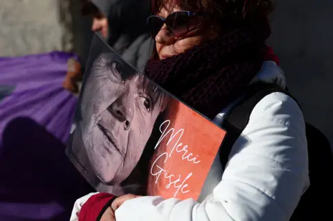 Reuters A woman wearing a white coat and sunglasses holds a sign with Gisèle Pelicot's face with the words 