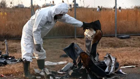 EPA A member of Korea Crime Scene Investigation in a white hazmat suit inspects the wreckage of the Jeju Air aircraft at Muan International Airport. They are holding up what appears to be the remains of a plane seat