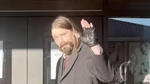 PA Media Benjamin Clarke is waving as he walks out of the court building. He is wearing black fingerless gloves and a long, grey coat. He has long brown hair and a beard.