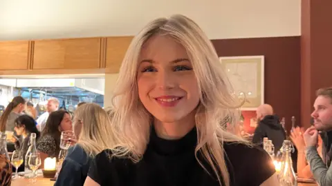 Evie Summers Evie Summers, who has blonde hair, smiling in a restaurant, wearing a black top