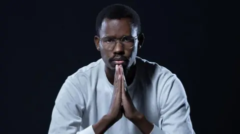 AFP Nelson Amenya, in a white top, holds his hands in front of him in a prayer position as he stares into the camera.